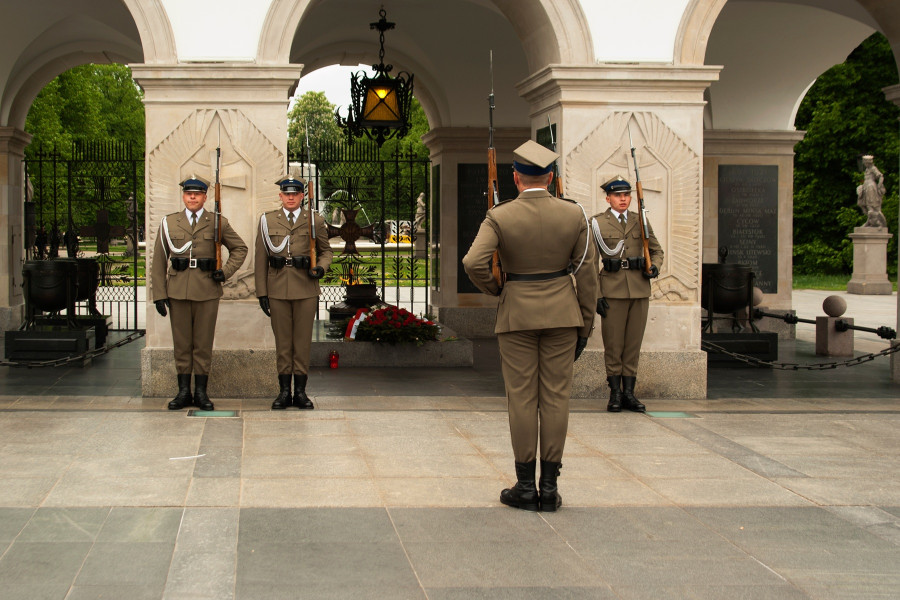Żołnierze Pułku Reprezentacyjnego Wojska Polskiego w pełnym umundurowaniu galowym, trzymający karabiny z bagnetami, pełnią wartę honorową przed pomnikiem na placu Piłsudskiego.